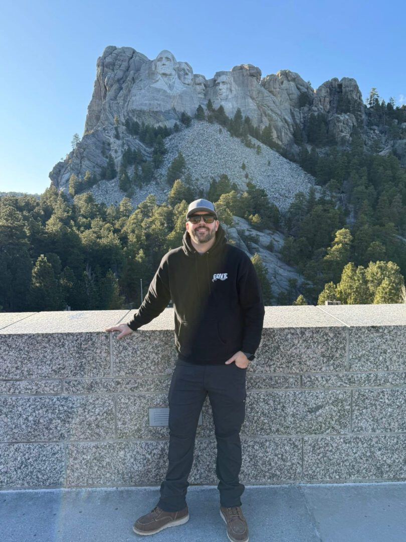 Man posing by a stone railing with a mountain backdrop.